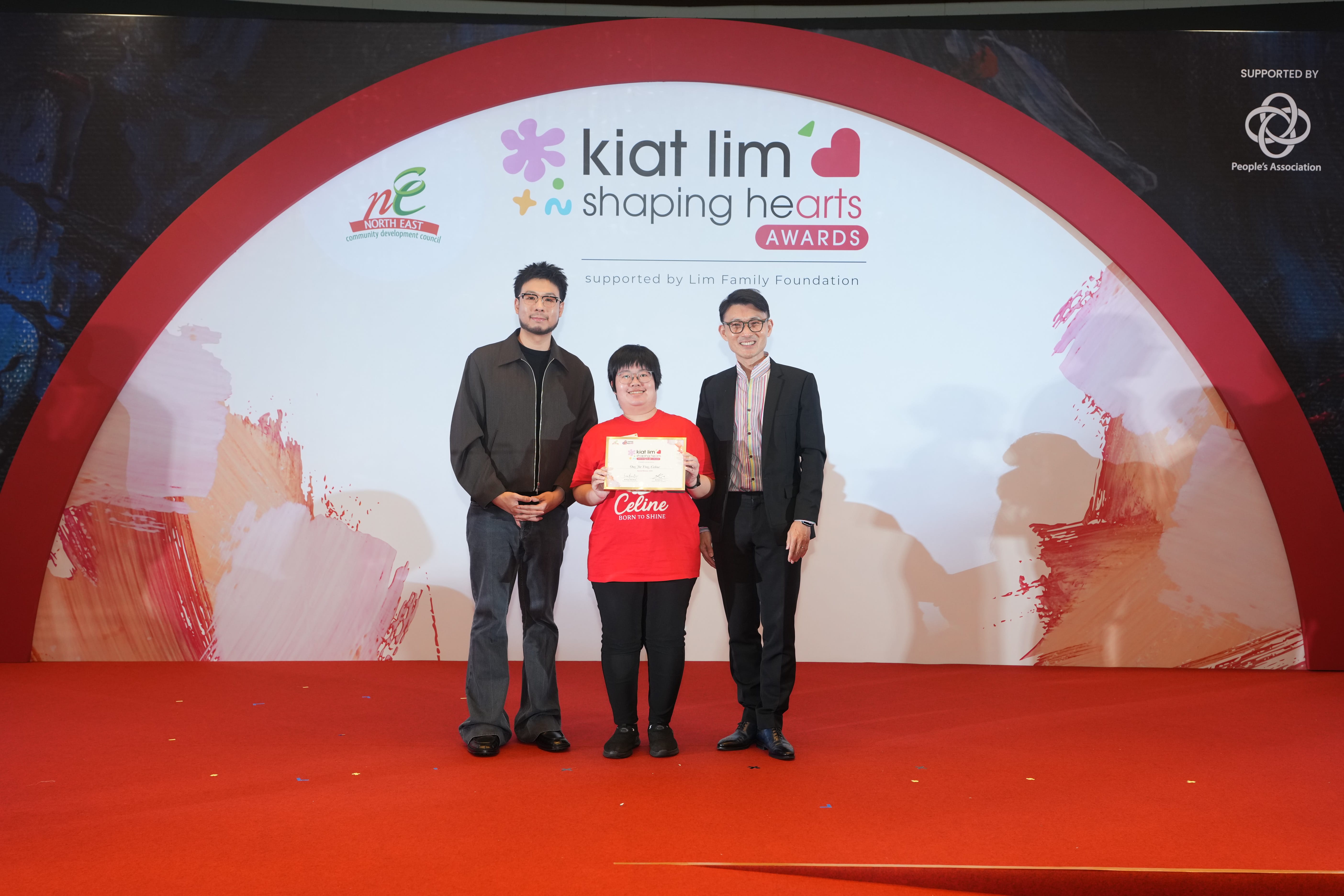 Award recipient standing on stage with the presenter, holding their certificate and posing for a group photo during the award ceremony, with a backdrop reading ‘Kiat Lim-Shaping Hearts Awards'.